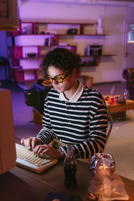 A young man with glasses types on a vintage computer in a cozy room lit by warm lighting.